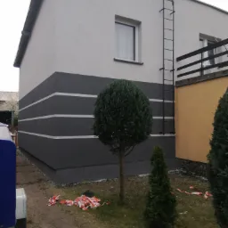 Dwukondygnacyjny budynek z elewacją w kolorach szarości, z poziomymi pasami. Na ścianie przymocowana drabina. Na trawniku porozrzucana taśma ostrzegawcza, sugerująca niedawno zakończone prace.