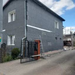 Dom w trakcie ocieplania szarym styropianem, widoczne fragmenty z nałożoną siatką i klejem, drabina oparta o ścianę, brama wjazdowa z czarnym płotem, pracownik w tle.