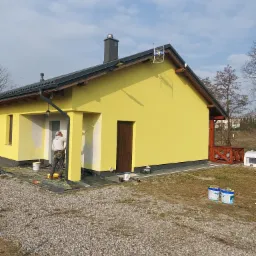 Malowanie fragmentu żółtej elewacji domu jednorodzinnego przez malarza z użyciem wałka, widoczne puszki z farbą, podłoże wysypane drobnym kamieniem.