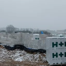 Wznoszenie ścian fundamentowych z bloczków betonowych H+H w zimowej scenerii; widoczny zarys budynku w surowym stanie, ośnieżone pole w tle i lekko prószący śnieg na bloczkach.