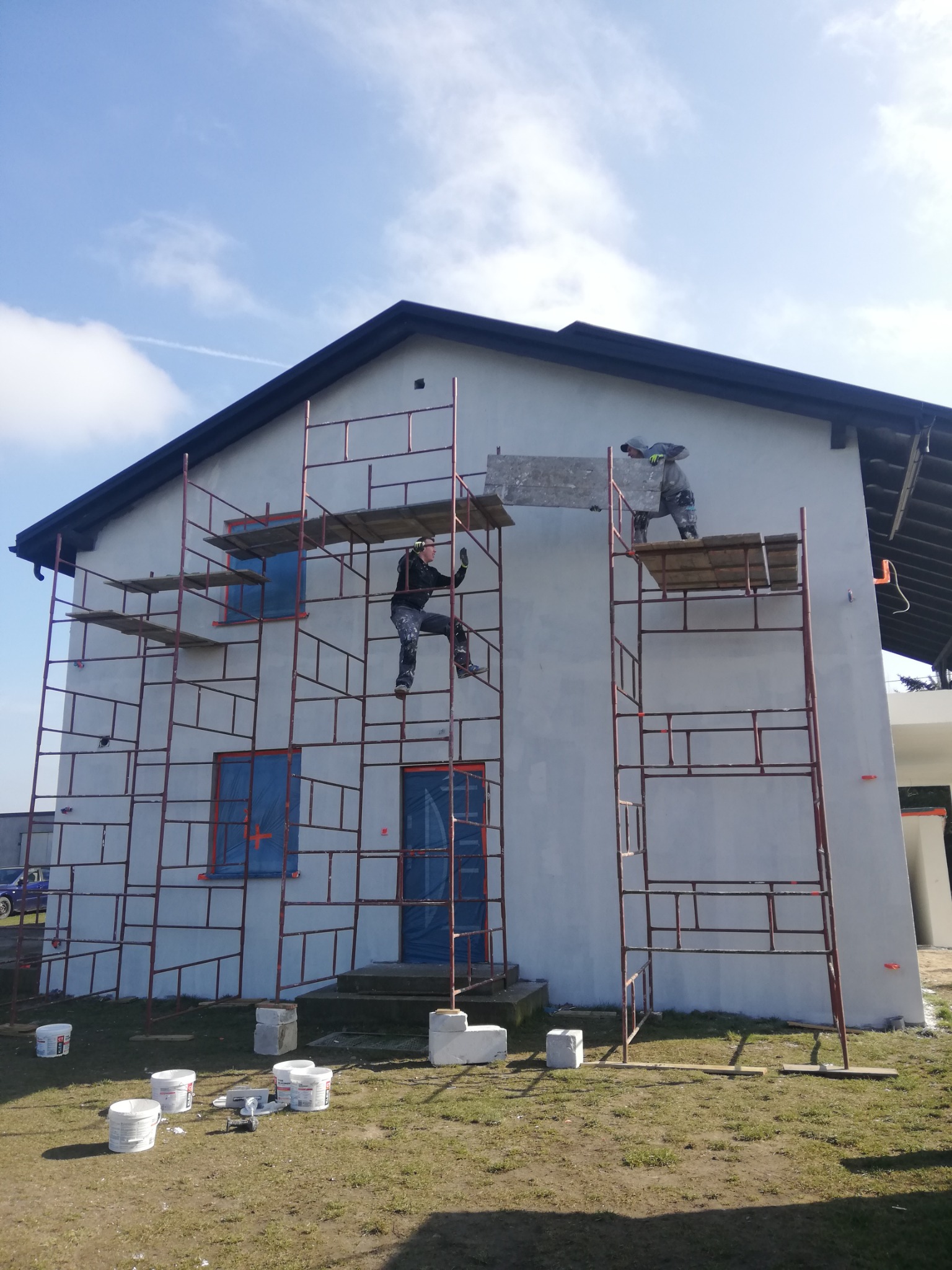 Dwóch pracowników na rusztowaniu przy białym domu z ciemnym dachem, okna zabezpieczone folią, na trawniku wiaderka z farbą i narzędzia.