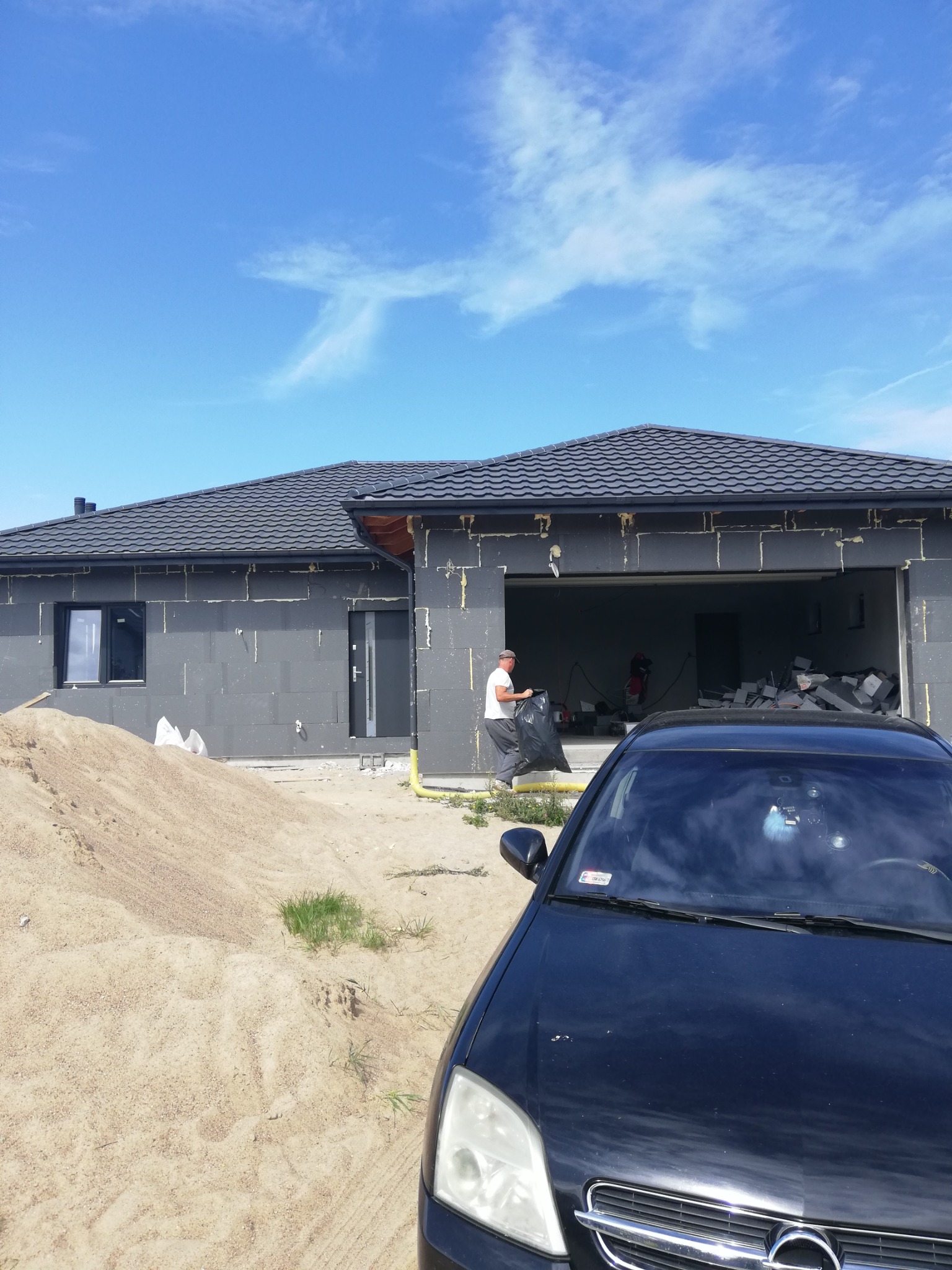 Dom w trakcie ocieplania styropianem grafitowym, widoczny pracownik z workiem na odpady, na pierwszym planie zaparkowany czarny samochód.