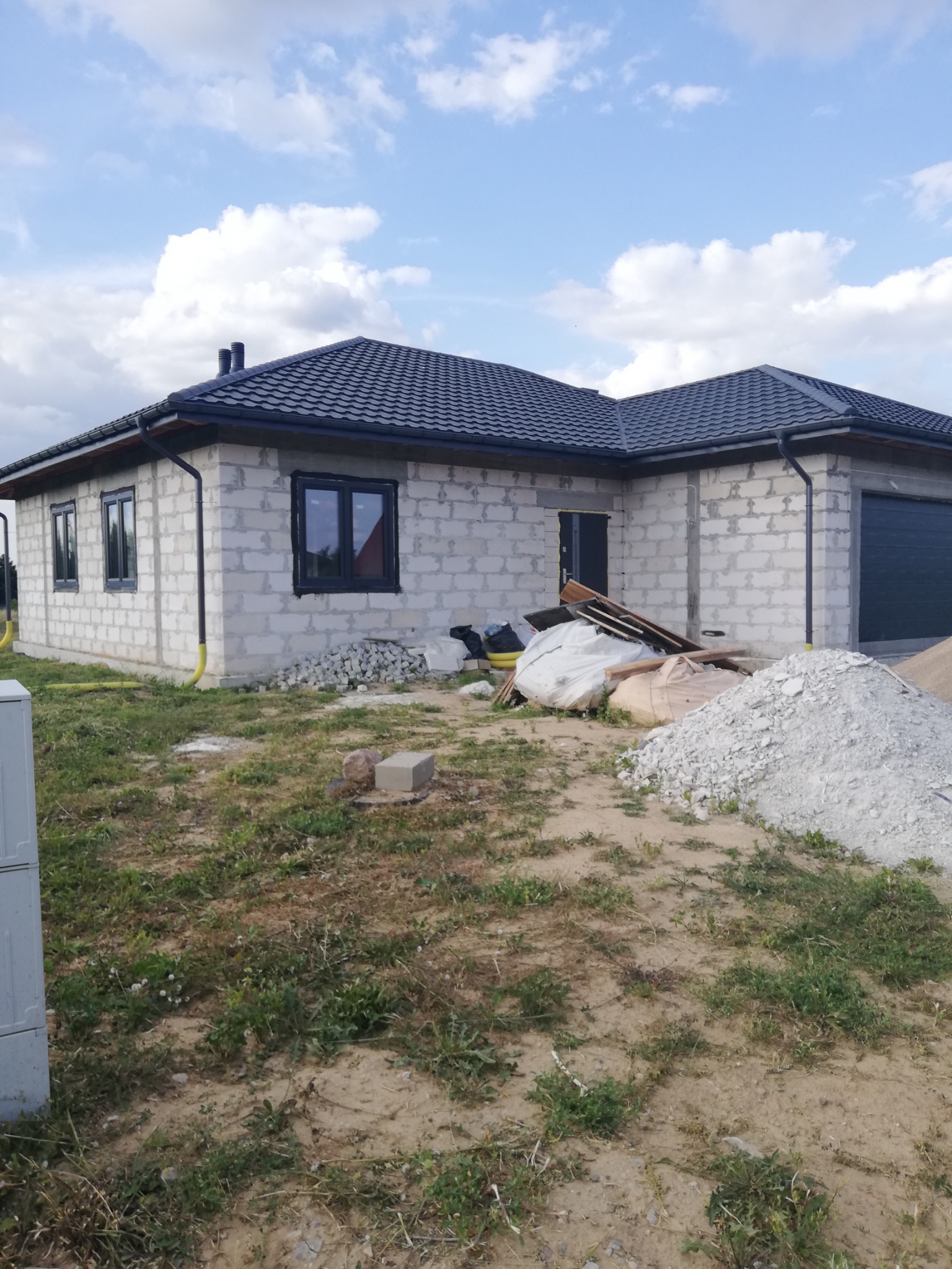 Surowy, parterowy dom z bloczków, z ciemną dachówką i orynnowaniem, w trakcie budowy, z widocznymi materiałami budowlanymi na działce.