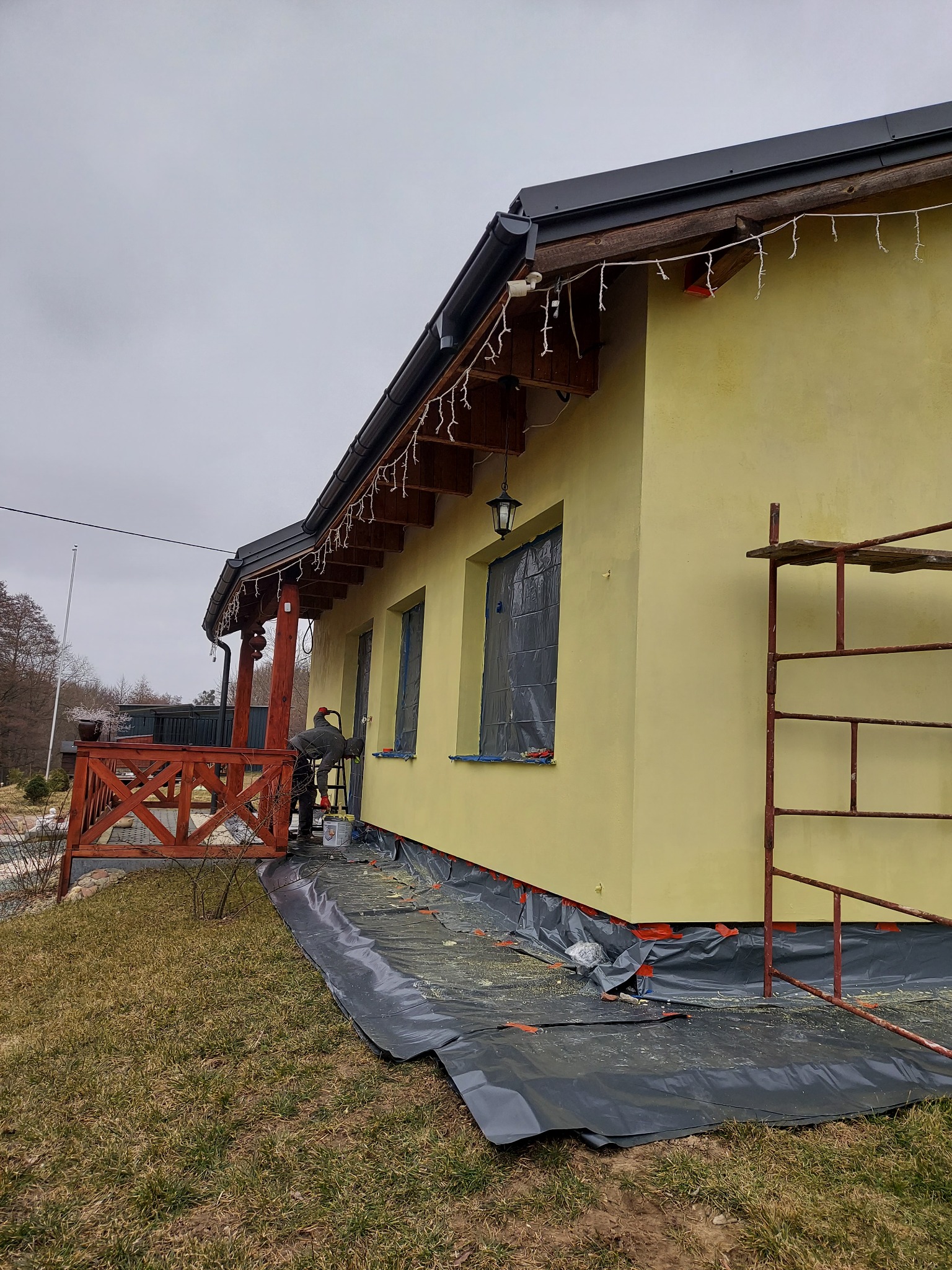 Żółta elewacja domu jednorodzinnego w trakcie malowania, okna zabezpieczone folią, widoczne rusztowanie i rozłożona folia ochronna na trawniku.