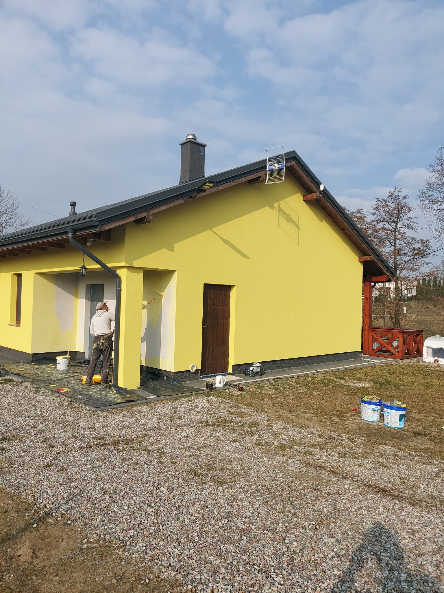 Malowanie fragmentu żółtej elewacji domu jednorodzinnego przez malarza z użyciem wałka, widoczne puszki z farbą, podłoże wysypane drobnym kamieniem.