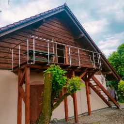 Drewniany budynek z balkonem z balustradą nierdzewną i schodami zewnętrznymi, widok z dołu na tle pochmurnego nieba.