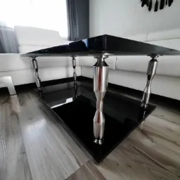 Nowoczesny stolik kawowy z czarnym, błyszczącym blatem i srebrnymi, metalowymi nogami o nietypowym kształcie, ustawiony na drewnianej podłodze w salonie z białą sofą w tle.