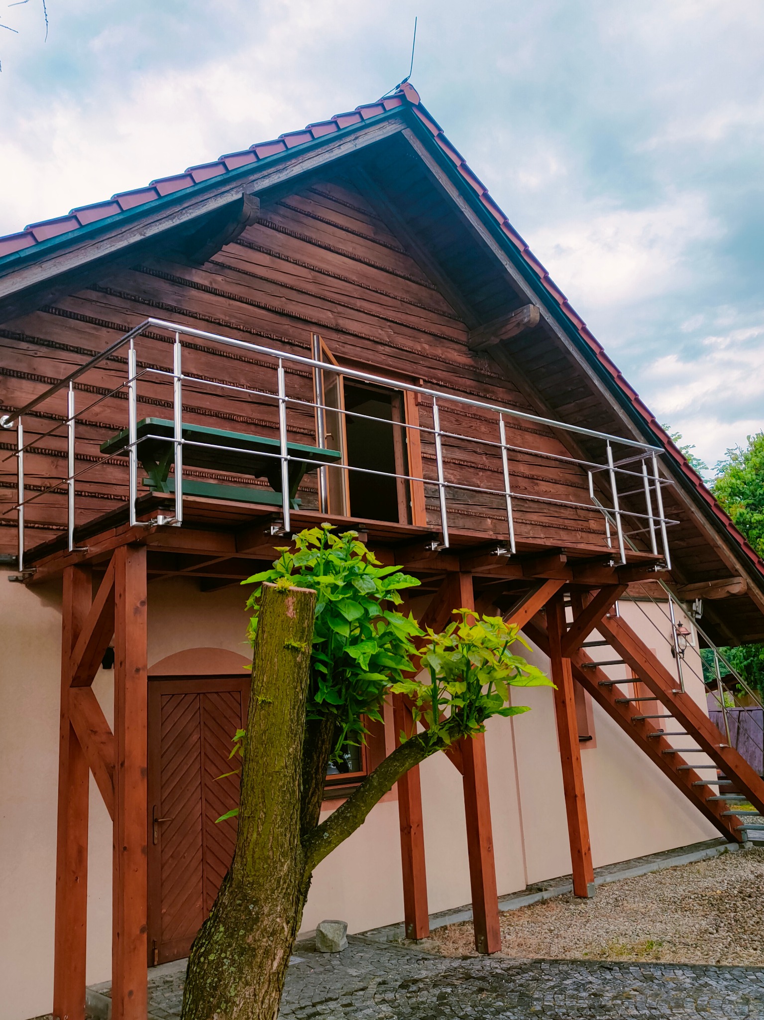 Drewniany dom z balkonem zabezpieczonym stalową, nierdzewną barierką, widoczne schody prowadzące na balkon i zielona ławka.