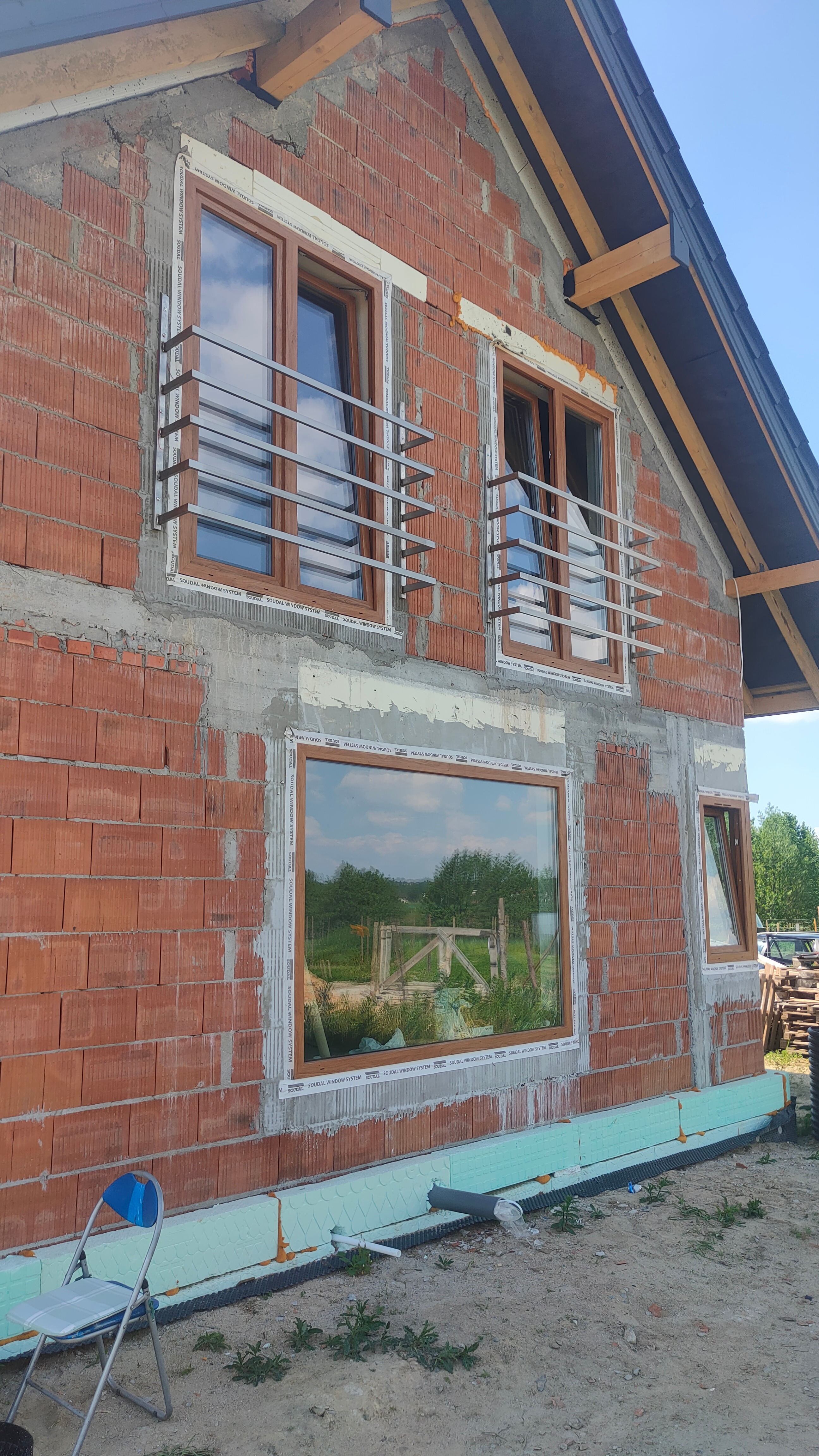 Surowy stan budynku w trakcie budowy z zamontowanymi oknami z drewnianą ramą i prostymi, poziomymi balustradami ze stali nierdzewnej na dwóch oknach, widoczne ocieplenie fundamentów z niebieskiego...