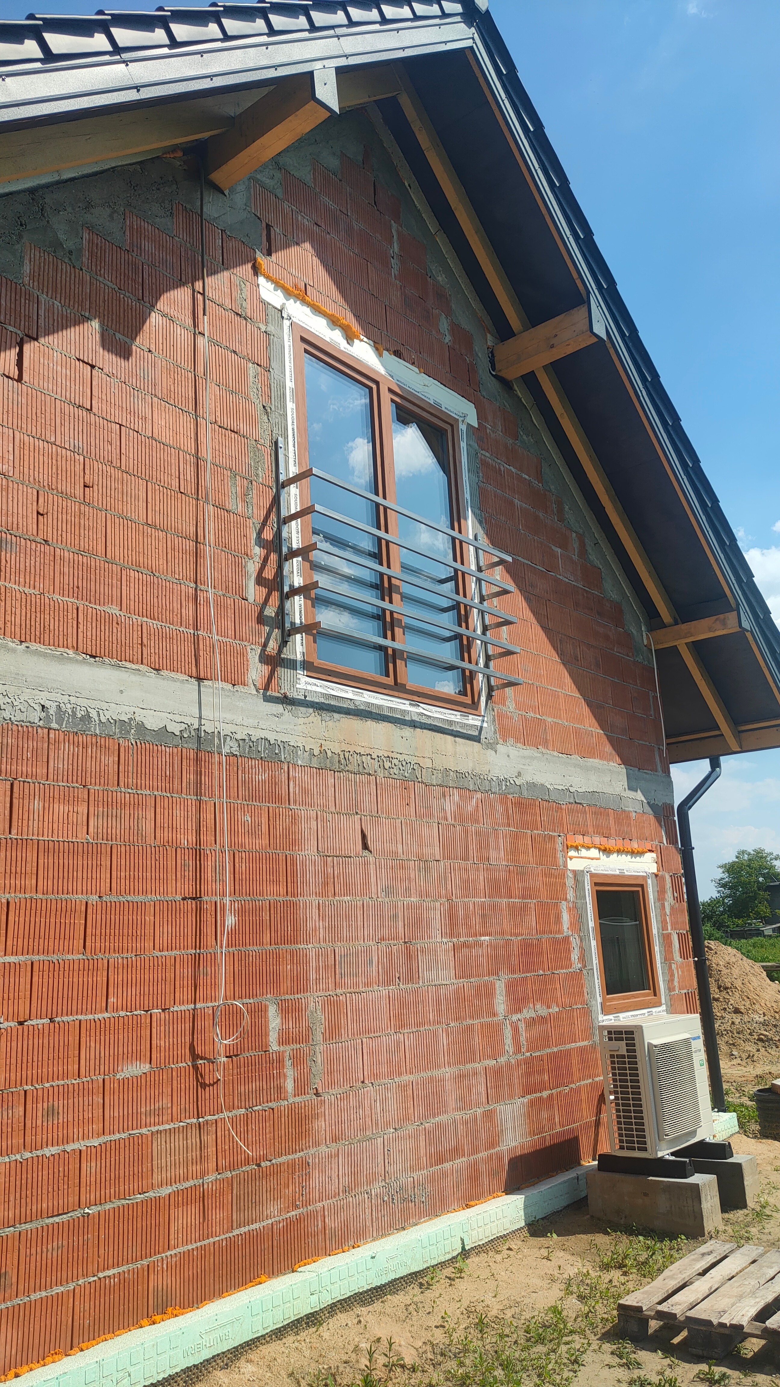 Surowa ściana budynku z czerwonej cegły z zamontowanym oknem z brązową ramą i prostą, poziomą, metalową konstrukcją zabezpieczającą, prawdopodobnie w trakcie montażu.