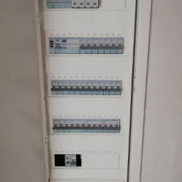 Otwarta skrzynka elektryczna z bezpiecznikami i wyłącznikami różnicowoprądowymi marki Hager, rozmieszczonymi w rzędach od 1 do 60, zamontowana na białej ścianie.