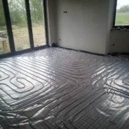 Instalacja ogrzewania podłogowego w nowym budynku, rury grzewcze ułożone na folii z siatką w pomieszczeniu z dużymi oknami tarasowymi.