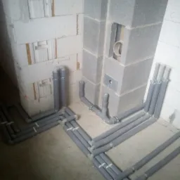 Szare rury instalacyjne w izolacji, prowadzone wzdłuż ścian z bloczków betonowych i betonu komórkowego na surowym etapie budowy.