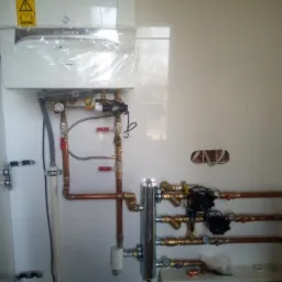 Nowy, biały piec gazowy De Dietrich, jeszcze w folii ochronnej, zamontowany na ścianie z białych płytek. Widoczne miedziane rury instalacji oraz liczne zawory i manometry. Część ściany po prawej...