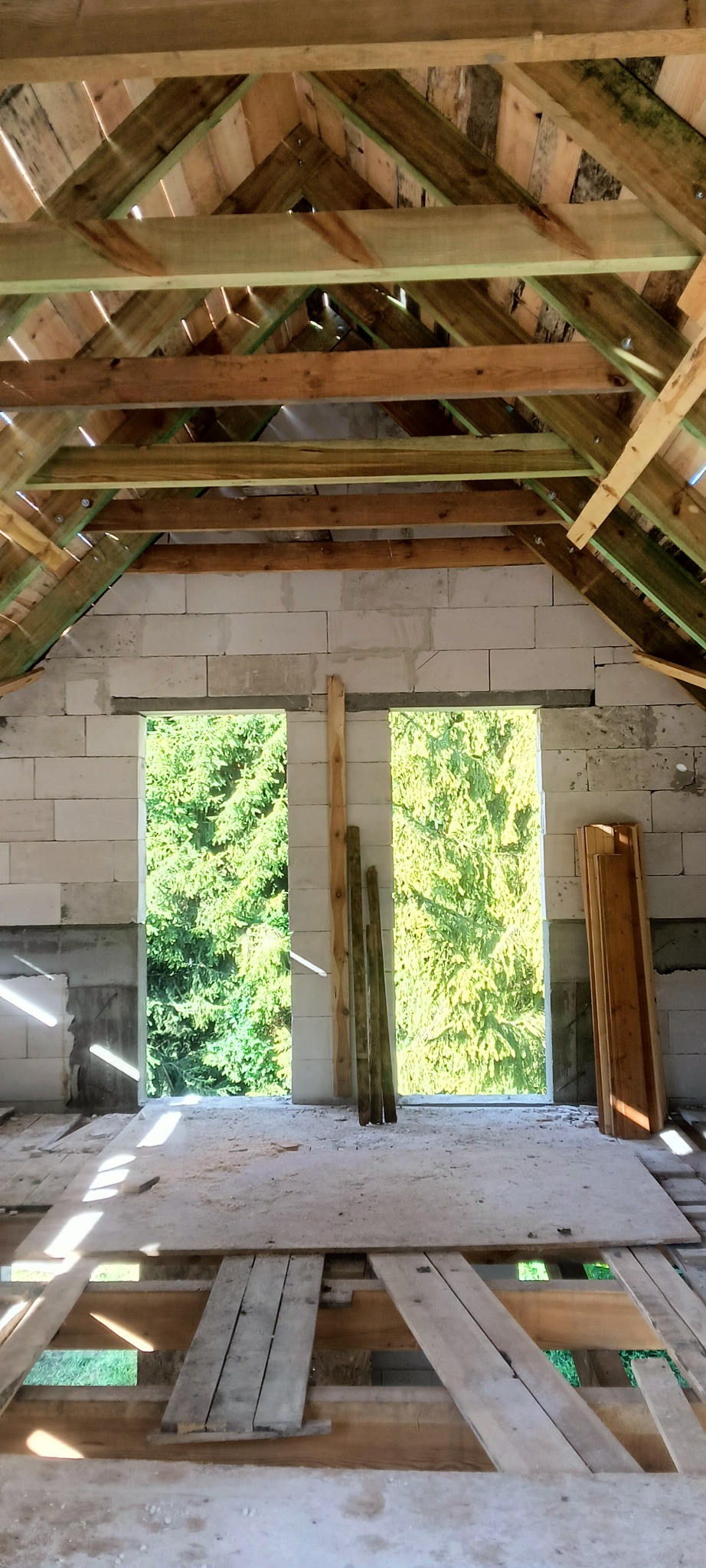 Surowy stan domu w trakcie budowy, widoczne ściany z bloczków, drewniana konstrukcja dachu z zielonymi impregnatami, deski podłogowe i widok na drzewa przez otwory okienne.