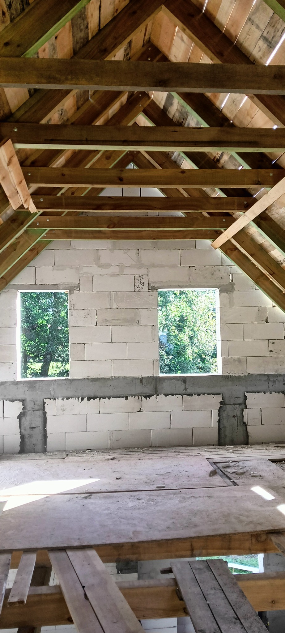 Wnętrze domu w trakcie budowy: surowe ściany z betonu komórkowego z otworami okiennymi, widoczna konstrukcja drewniana dachu z deskowaniem, częściowo ułożona podłoga z desek. Przez okna widoczne...