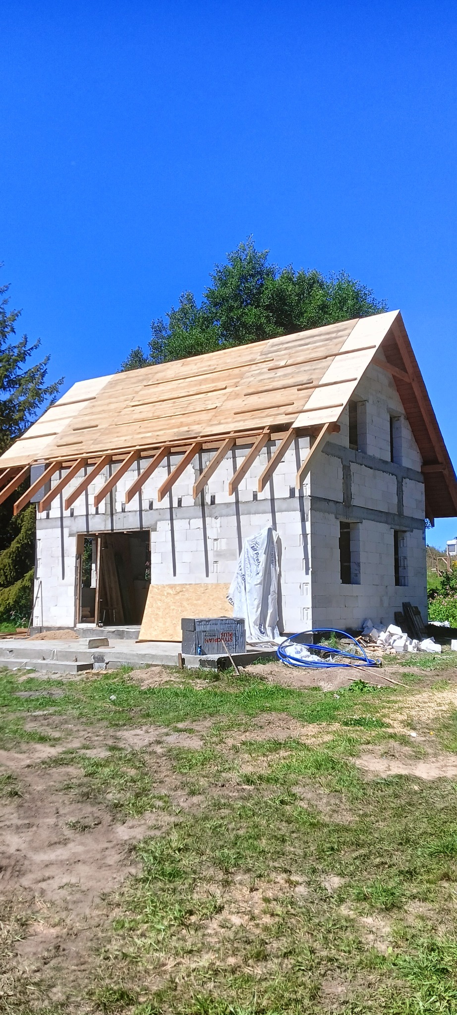 Budowa domu murowanego w trakcie realizacji: ściany z bloczków, częściowo wykonany drewniany szkielet dachu, materiały budowlane na posesji, błękitne niebo.