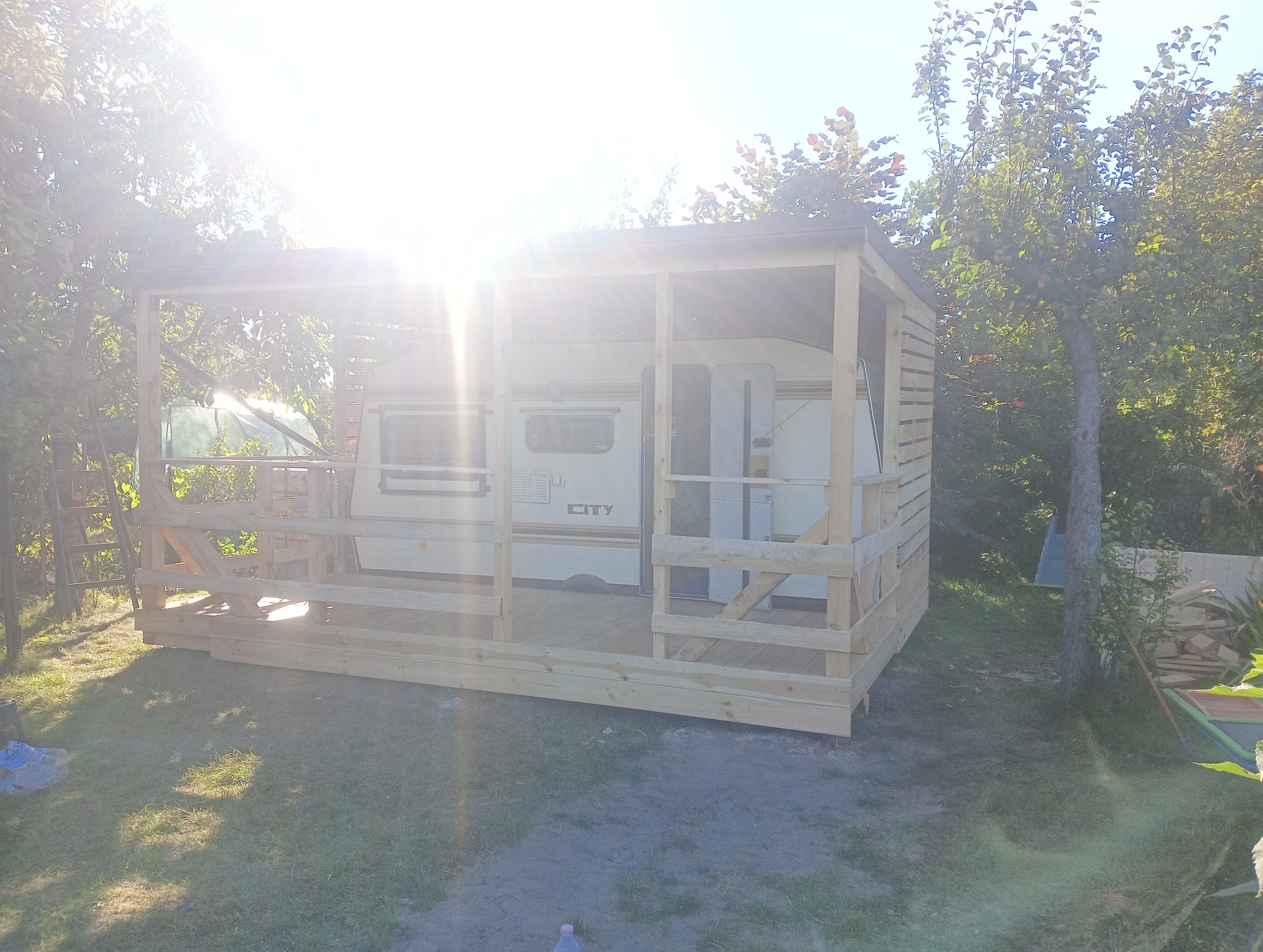 Drewniana konstrukcja zadaszenia tarasu w trakcie budowy, częściowo osłaniająca przyczepę kempingową na działce, widoczne elementy konstrukcyjne i deski w jasnym drewnie, zieleń w tle.