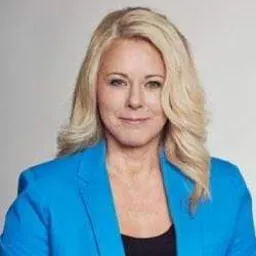 Portret uśmiechniętej blondynki w niebieskiej marynarce na jasnoszarym tle, sugerujący profesjonalizm i pewność siebie.