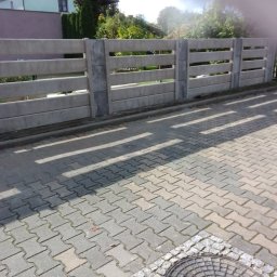 Dominika Pawłowska - Betonowe ogrodzenie panelowe z trzema poziomymi przęsłami, rzucające cienie na brukowaną nawierzchnię. W tle widoczny dom i zieleń.