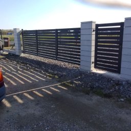 Dominika Pawłowska - Nowoczesne, poziome ogrodzenie panelowe w kolorze antracytowym z bramką, osadzone na betonowych słupkach. Widoczne cienie rzucane przez panele na żwirową nawierzchnię.