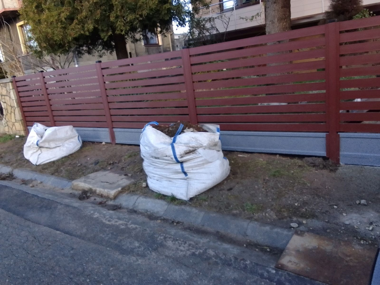 Nowoczesne ogrodzenie panelowe w kolorze brązowym z poziomymi szczeblami, widoczne worki z ziemią po montażu, ujęcie z poziomu ulicy.