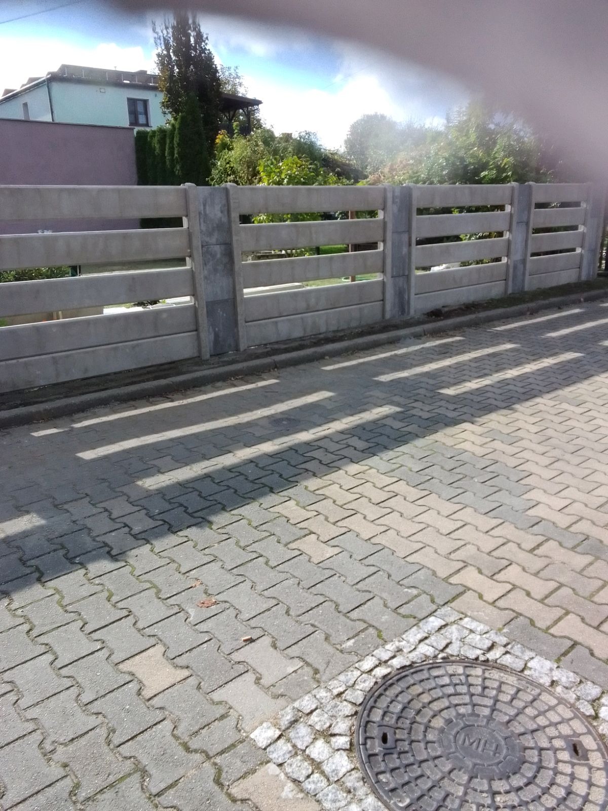 Betonowe ogrodzenie panelowe z trzema poziomymi przęsłami, rzucające cienie na brukowaną nawierzchnię. W tle widoczny dom i zieleń.