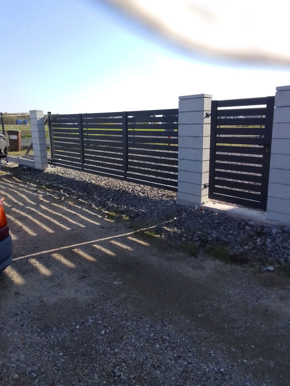 Nowoczesne, poziome ogrodzenie panelowe w kolorze antracytowym z bramką, osadzone na betonowych słupkach. Widoczne cienie rzucane przez panele na żwirową nawierzchnię.