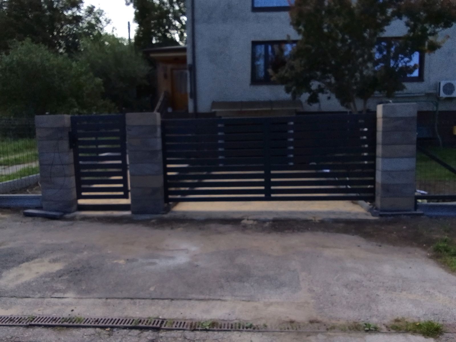 Nowoczesna, pozioma brama wjazdowa z metalowymi przęsłami i kamiennymi słupkami, na tle domu jednorodzinnego. Widoczne detale wykończenia i otoczenie.