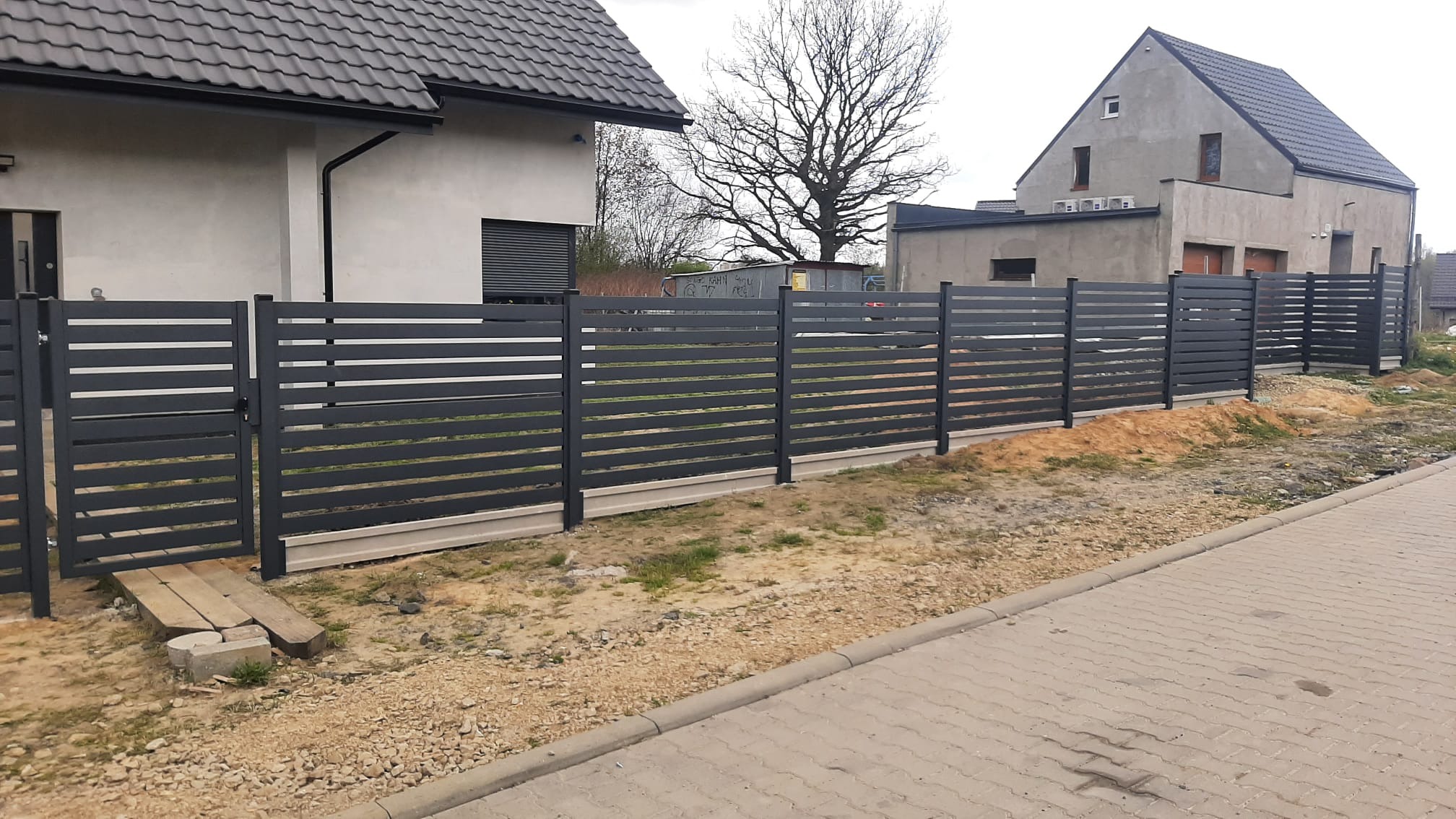 Nowoczesne, poziome ogrodzenie panelowe w kolorze grafitowym, z furtką i betonową podmurówką, na tle domu jednorodzinnego i chodnika z kostki.