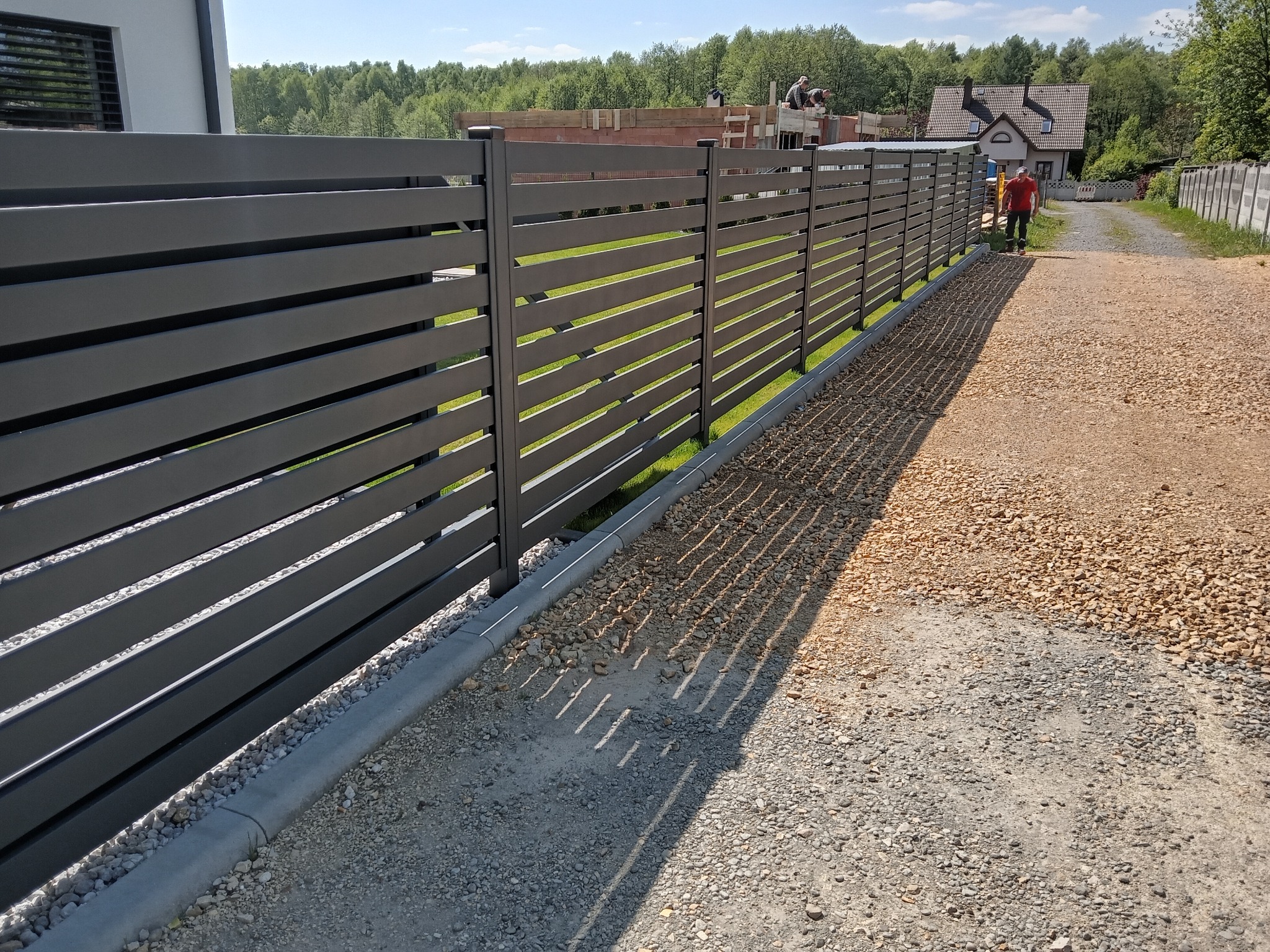 Nowoczesne, poziome ogrodzenie panelowe w kolorze grafitowym, z widocznym fundamentem i ścieżką z kamienia. W tle budowa domu i zieleń drzew.
