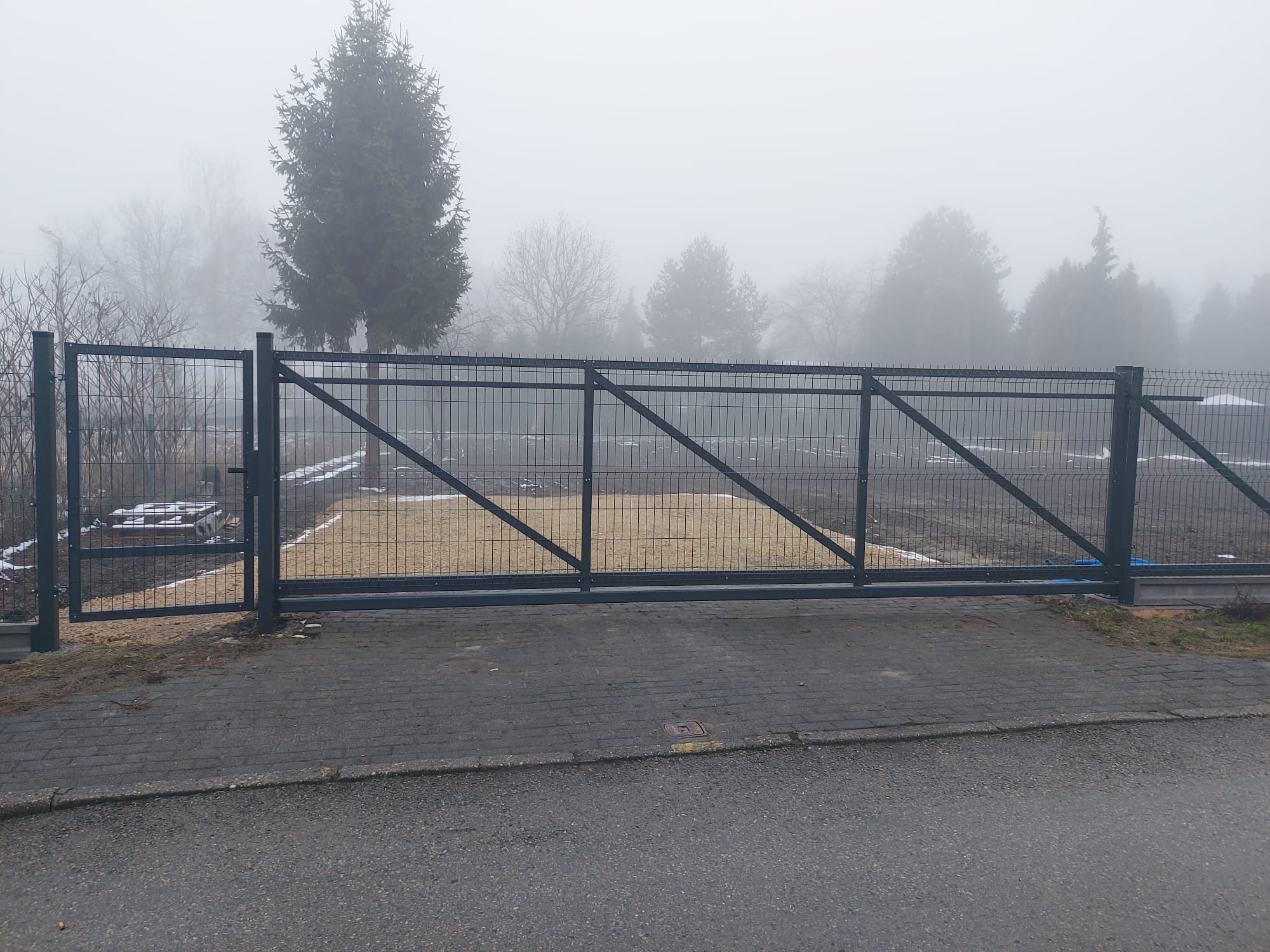 Nowoczesna, przesuwna brama panelowa w kolorze grafitowym, z widoczną konstrukcją wzmacniającą, na tle mglistego krajobrazu. Kostka brukowa przed bramą.