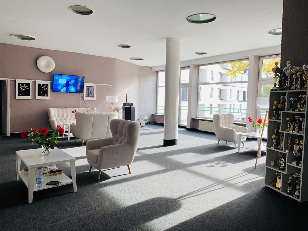 Jasne wnętrze z sofami, fotelami i regałem z trofeami, prawdopodobnie poczekalnia lub strefa relaksu, z widocznymi promieniami słonecznymi na podłodze.