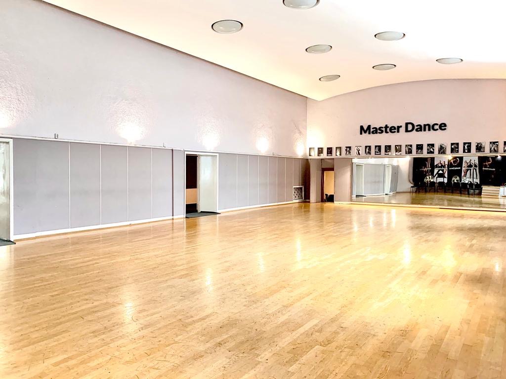 Pusta sala taneczna z drewnianą podłogą, lustrami na ścianie, plakietkami na górze i napisem Master Dance na ścianie, oświetlona okrągłymi lampami sufitowymi.