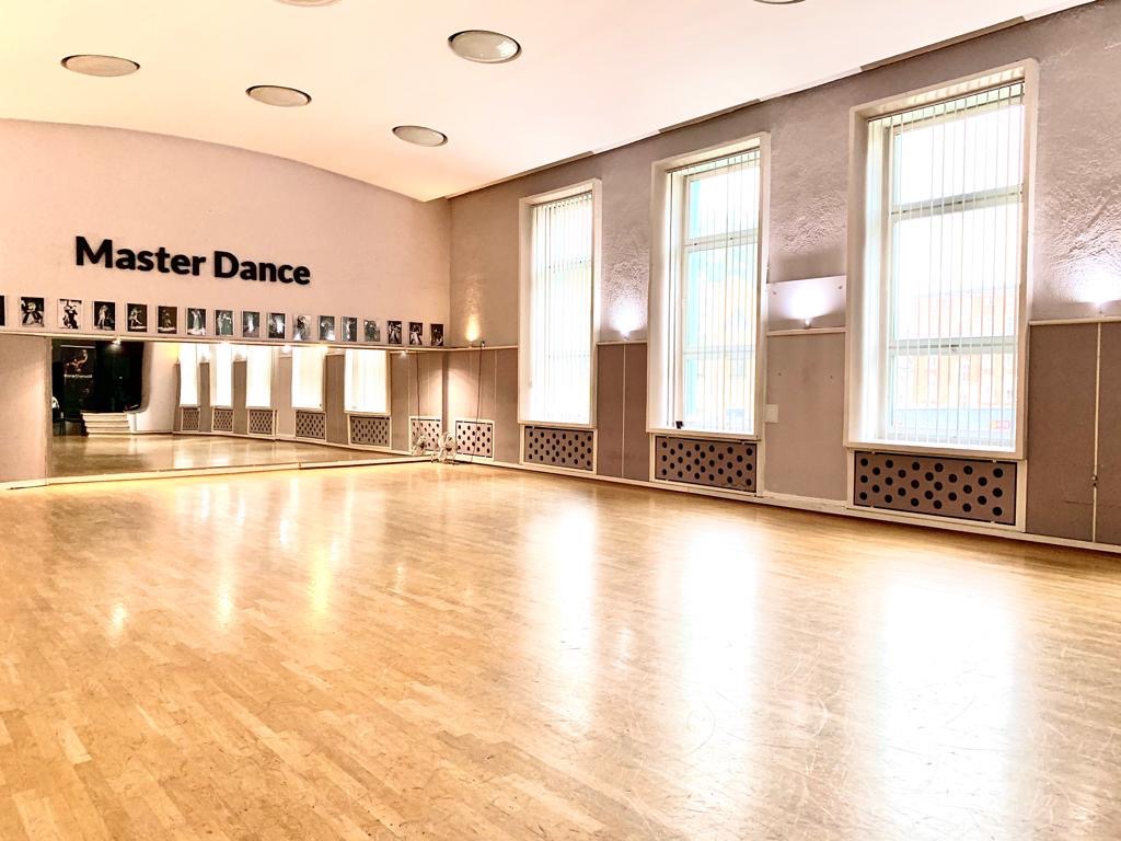 Pusta sala z lustrami i parkietem, logo 'Master Dance' na ścianie, rzędy zdjęć pod sufitem, trzy okna z żaluzjami.