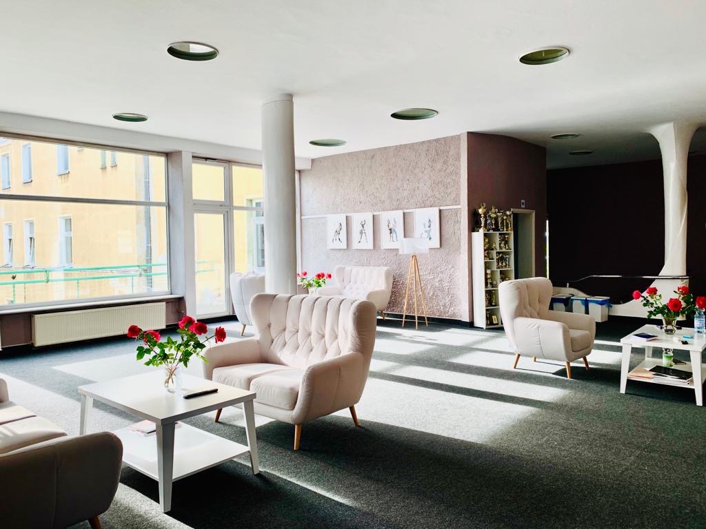 Jasne wnętrze poczekalni z sofami w odcieniach beżu, stolikami z bukietami czerwonych róż, widok na trofea w regale i grafiki taneczne na ścianie.