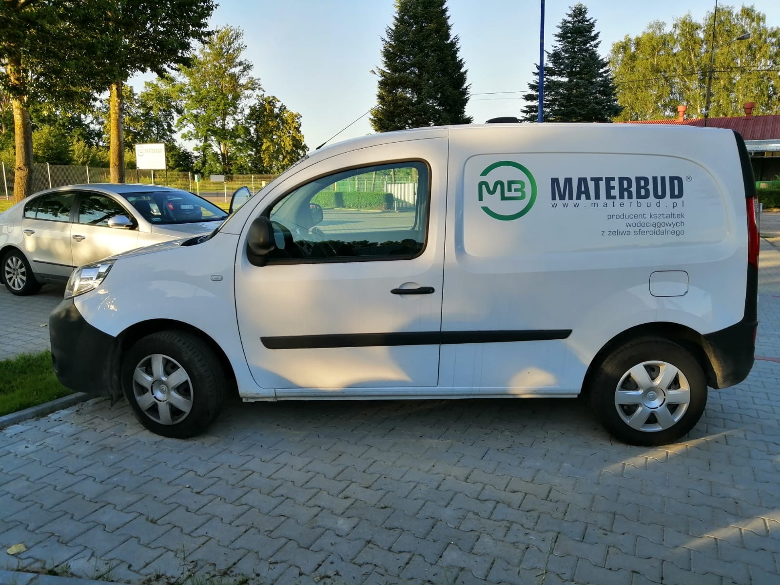 Biały samochód dostawczy z reklamowym nadrukiem logo firmy Materbud i danymi kontaktowymi, zaparkowany na kostce brukowej, w tle drzewa i zaparkowany samochód osobowy.