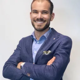 Uśmiechnięty mężczyzna w niebieskiej marynarce i koszuli, z poszetką w kieszonce, pozuje z założonymi rękami na jasnym tle.