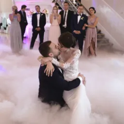 Pocałunek pary młodej w gęstej mgle podczas wesela, goście obserwują z tyłu, panuje uroczysta atmosfera.