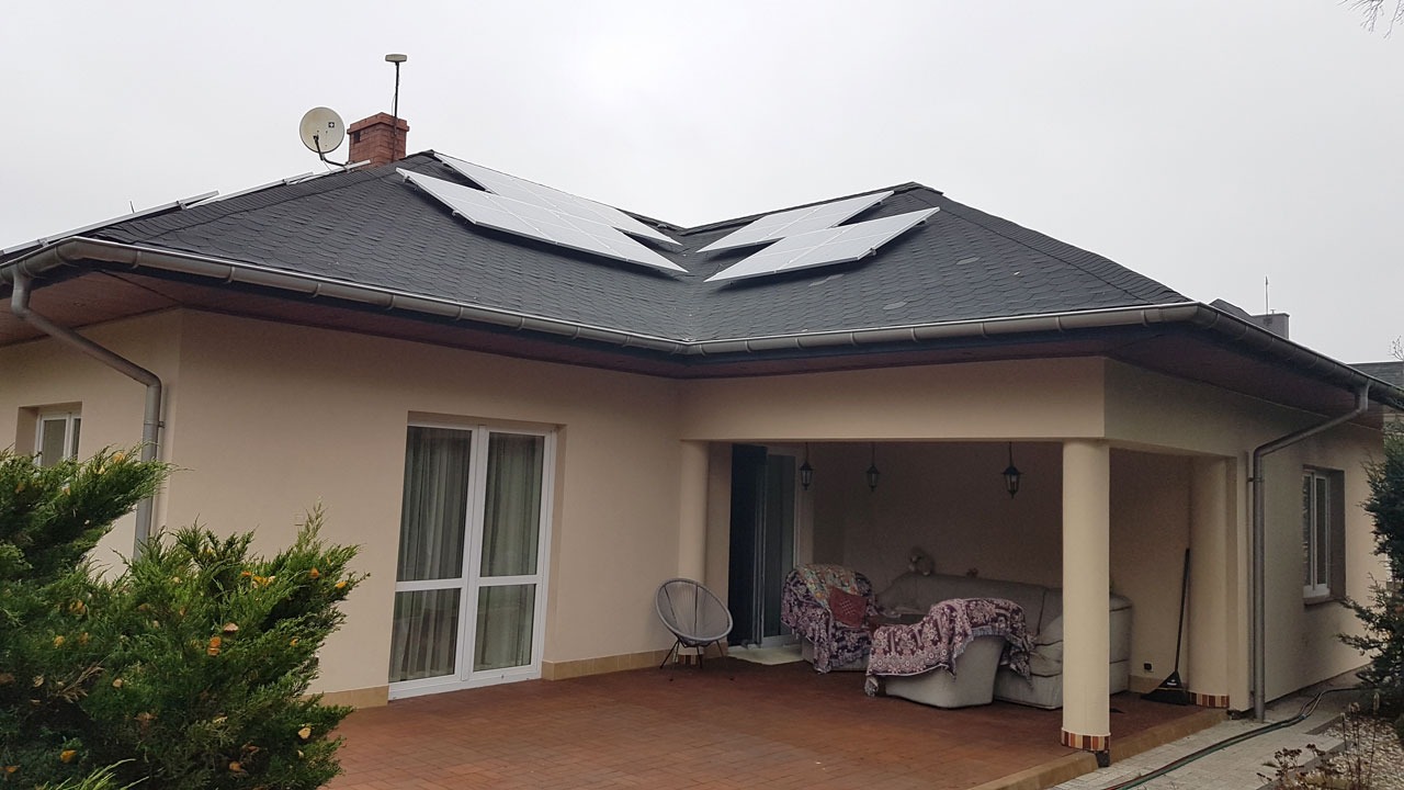 Dom jednorodzinny z zamontowanymi panelami fotowoltaicznymi na dachu pokrytym czarną dachówką bitumiczną, widoczne również antena satelitarna i komin.