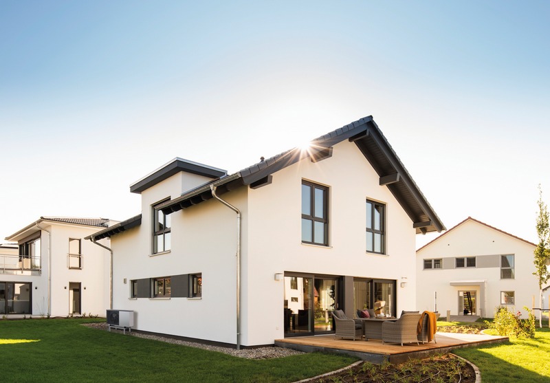Nowoczesny dom jednorodzinny z białymi ścianami, ciemną stolarką okienną i dachem, z tarasem i meblami ogrodowymi na tle zielonego trawnika i błękitnego nieba z promieniem słońca.