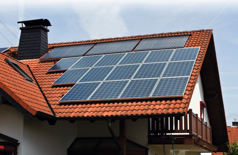 Panele słoneczne i kolektory słoneczne zamontowane na dachu z czerwonej dachówki, widoczne okno dachowe i komin z ciemnej cegły.