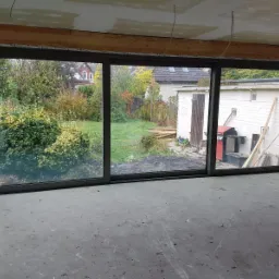 Duże, przesuwne okno z ciemną ramą, widok na ogród z domem i budynkiem gospodarczym, surowy stan wykończenia wnętrza.