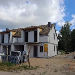 Surowy stan bliźniaka z białego betonu komórkowego w trakcie budowy, widoczne drewniane krokwie dachu i palety z materiałami budowlanymi.