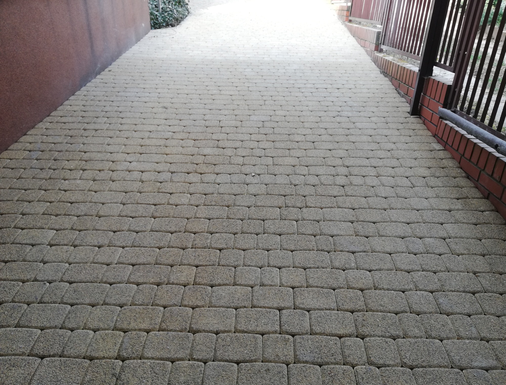Ułożona szara kostka brukowa tworząca wjazd, widoczna ściana budynku z lewej strony i ogrodzenie z prawej strony.