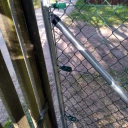 Fragment zielonej siatki ogrodzeniowej, połączonej z drewnianym płotem za pomocą metalowych słupków i zielonych uchwytów