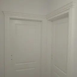 Dwa białe, klasyczne drzwi wewnętrzne z ozdobnymi ościeżnicami, widoczne pod kątem w rogu pomieszczenia, z widocznymi zawiasami.