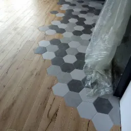 Przejście pomiędzy drewnianą podłogą a heksagonalnymi płytkami w odcieniach szarości, częściowo przykryte folią ochronną, widok na fragment grzejnika i szafki kuchennej.