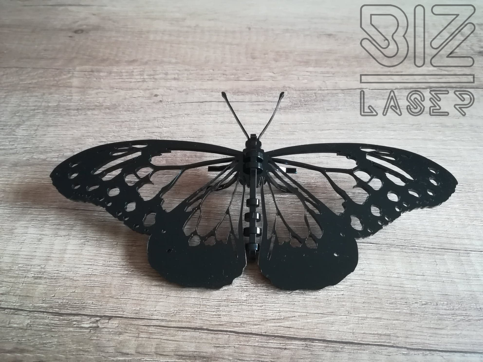 Czarny, ażurowy motyl wykonany techniką laserową, umieszczony na drewnianym tle, z widocznym logo firmy BIZ LASER w tle.
