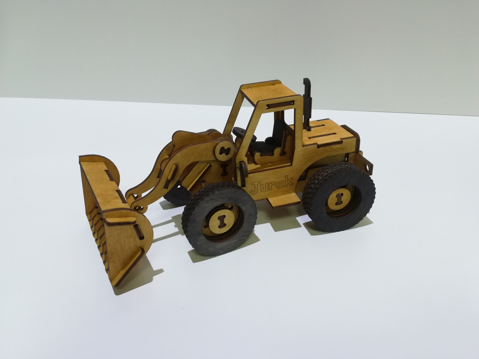 Model drewnianego traktora z ruchomą łyżką, z napisem 'Jurek' na boku, na białym tle.